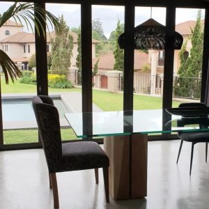 Folding aluminium doors open to a bright garden and pool area from a dark modern dining room with glass table and chairs.