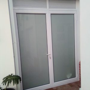 Frosted aluminium french doors with white frame and top air vent, installed in a small entry alcove with a potted plant.