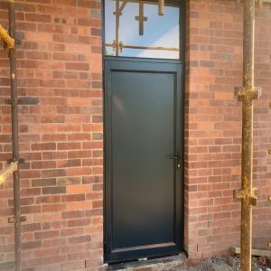 Black aluminium single hinge door with top window, fitted in a red brick wall with scaffolding on both sides.
