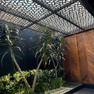 Geometric-patterned aluminium pergola providing partial shade over garden area with tropical plants and wooden panel wall.