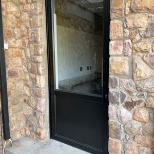 Black aluminium single hinge door set in a natural stone wall, showing a partial interior view through the glass panel.